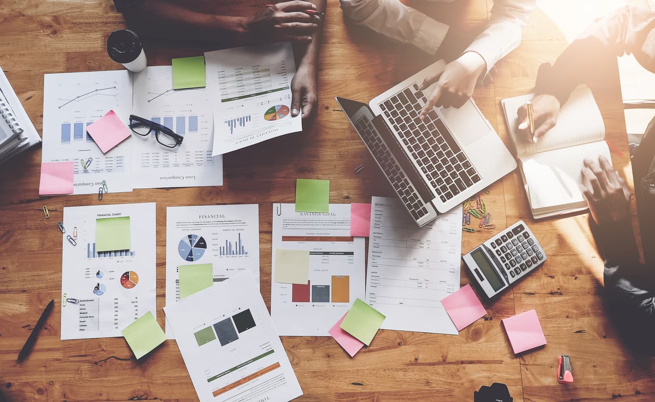 Overhead view of a team working at a wooden table with laptops, financial charts, sticky notes and stationery, symbolizing collaboration, planning and data analysis.