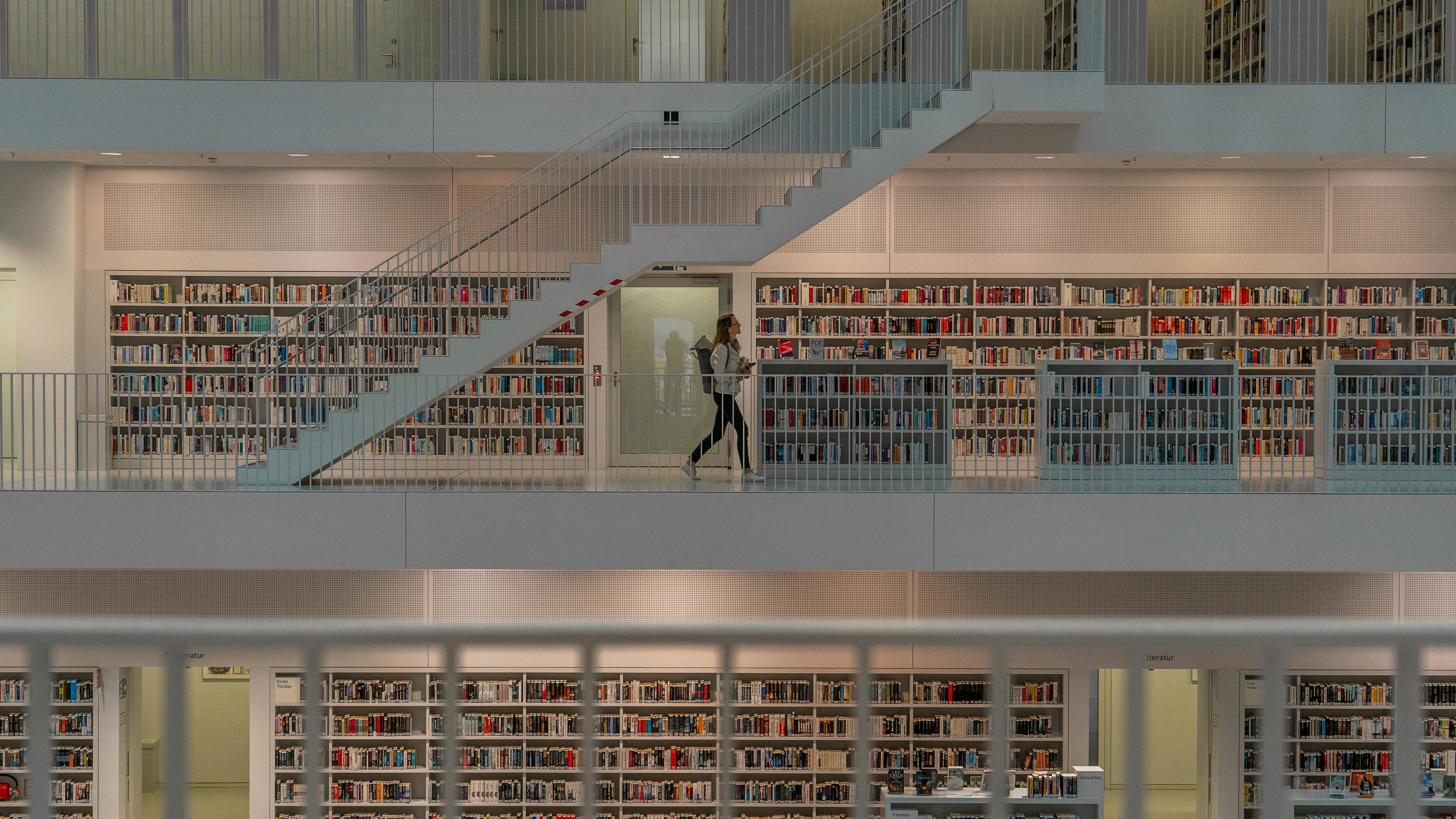 A person walking along an upper level of a spacious, multi-story library with shelves filled with books and a prominent staircase in the center